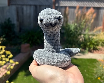 May include: A gray crocheted snake plush toy with black eyes and a smiling mouth. The toy is being held in a person's hand.