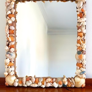 May include: Square mirror with a frame completely covered in various seashells. The shells are in shades of white, beige, orange, and brown, creating a textured border around the reflective glass. The mirror is a decorative coastal accent.