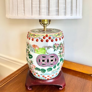 May include: A decorative ceramic lamp with a white pleated shade. The lamp base is a cylindrical shape with a colorful floral and landscape design, set on a dark red wooden base. The lamp has a brass neck.
