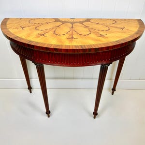 May include: A half-circle wooden table with a light-colored, patterned top and a dark red-brown apron. The table has four fluted legs and is set against a white wall. The top features a decorative inlay design.