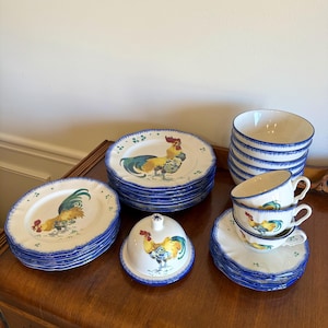 May include: A collection of white ceramic dinnerware with a rooster motif. The set includes plates, bowls, teacups, saucers, and a covered dish, all with blue rims. The rooster design features vibrant colors.