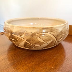 May include: A beige ceramic bowl with a wide, shallow design. The bowl features a decorative pattern of stylized leaves in shades of brown around the rim. The bowl is sitting on a wooden surface.