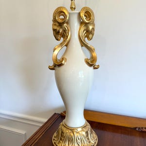 May include: A white ceramic table lamp with gold accents. The lamp features two gold ram head designs on the sides and a decorative gold base. The lamp has a brass neck and a standard light bulb socket.