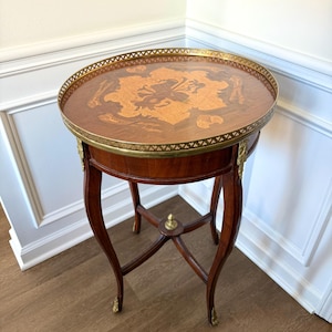 May include: A round, antique side table with a dark wood finish. The tabletop features a floral inlay design and a brass rim. The table has curved legs with brass accents and stands approximately 28 inches tall.