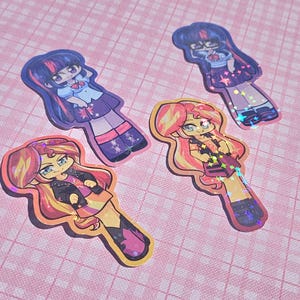 May include: Four colorful stickers featuring cartoon characters with long hair and stylish outfits. The stickers have a holographic effect and are placed on a pink checkered background. The characters have a cute, anime-inspired design.