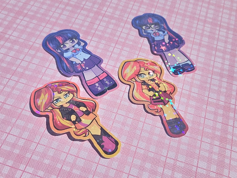 May include: Four colorful stickers featuring cartoon characters with long hair and stylish outfits. The stickers have a holographic effect and are placed on a pink checkered background. The characters have a cute, anime-inspired design.