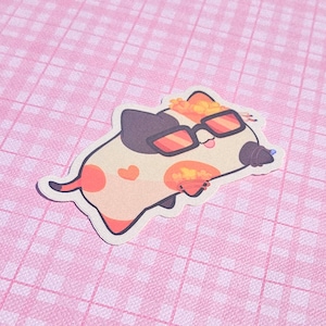 May include: A sticker featuring a cartoon cat with sunglasses. The cat has a cream-colored body with orange spots and a black and orange head. It is wearing orange-tinted sunglasses and has a small heart on its side. The sticker is on a pink checkered background.