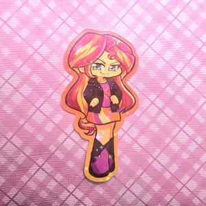 May include: A colorful sticker featuring a cartoon character with long, flowing hair in shades of yellow, orange, and pink. The character wears a black jacket, a pink shirt, and a skirt. The sticker has a glittery, holographic finish and is set against a pink background.