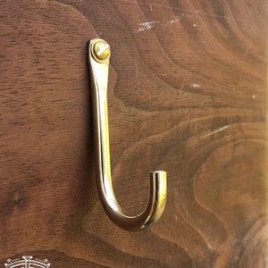 May include: A brass hook with a rounded end, mounted on a wooden surface.