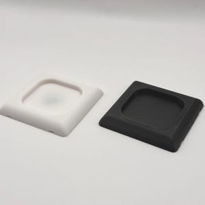 Può includere: Sono presentati due sottobicchieri quadrati, uno bianco e uno nero. Ogni sottobicchiere ha un centro quadrato incassato. I sottobicchieri sono realizzati in un materiale solido e hanno un bordo leggermente smussato.