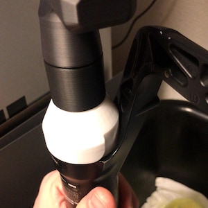 Puede incluir: Primer plano de un componente de suspensión de bicicleta en blanco y negro. El componente tiene una forma cilíndrica con una sección blanca y redondeada y una parte superior negra y angular. Una palanca negra está unida al lateral. La imagen está bien iluminada, resaltando las texturas y formas.