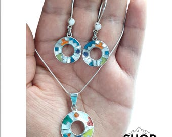 Può includere: Un set di collana e orecchini in argento con pendenti circolari con disegni geometrici colorati. Gli orecchini hanno una chiusura a gancio e una piccola perla bianca. Il ciondolo pende da una delicata catena.