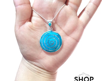 Può includere: Una collana d'argento con un pendente turchese. Il pendente è circolare con un design a spirale e una piccola pietra turchese in alto. Il logo Shop Peru si trova in basso a destra.