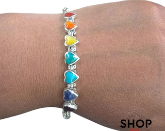Può includere: Un braccialetto d'argento con sette pietre a forma di cuore nei colori dell'arcobaleno: rosso, arancione, giallo, verde acqua, turchese, indaco e viola. Il braccialetto è mostrato su un polso.