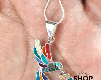 Può includere: Ciondolo a forma di colibrì in argento con un design a mosaico colorato. Le ali dell'uccello presentano dettagli rossi, arancioni, gialli, blu e verdi. Il colibrì tiene in mano un fiore con petali rosa, viola e verdi. Il ciondolo è appeso a una catena d'argento.