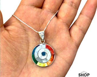 Può includere: Una collana con pendente in argento con un design circolare. Il pendente presenta un motivo a spirale con una gamma di colori: rosso, arancione, giallo, verde, blu e bianco. La collana è tenuta in una mano, con il testo "SHOP PERU" in basso a destra.