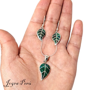 May include: A silver necklace and earrings set with green malachite leaf pendants. The jewelry is displayed on a hand.