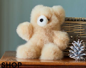 Può includere: Un orsacchiotto di alpaca marrone chiaro seduto su una superficie di legno. L'orso ha una faccia bianca con occhi neri e un naso nero.