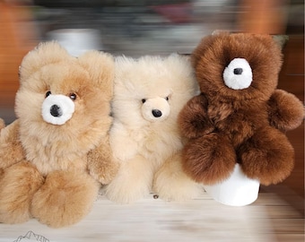 Può includere: Tre orsacchiotti di peluche in lana di alpaca. Gli orsi sono seduti su una superficie di legno. L'orso a sinistra è marrone chiaro, l'orso al centro è color crema e l'orso a destra è marrone.