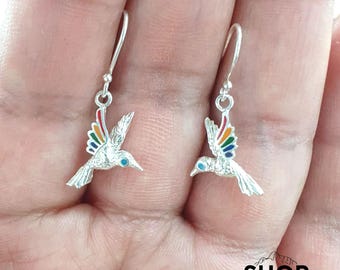 Può includere: Un paio di orecchini a forma di colibrì in argento. Ogni orecchino presenta un colibrì con ali color arcobaleno e un occhio blu. Gli orecchini hanno un semplice design a gancio. Le parole "SHOP" e "PERU" sono stampate in basso.