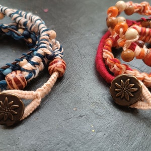 Könnte beinhalten: Zwei handgefertigte Armbänder mit bunten gewebten Fäden und Holzperlen. Jedes Armband hat einen Metallknopf mit einem Blumendesign.