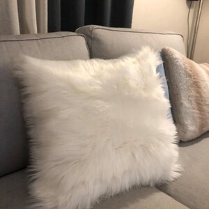 Puede incluir: Una almohada de piel sint&eacute;tica blanca sobre un sof&aacute; gris claro. La almohada es suave y esponjosa, y ser&iacute;a una gran adici&oacute;n a cualquier sala de estar.