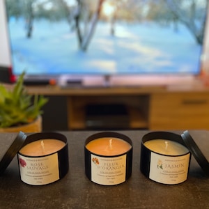 Peut inclure: Trois bougies en métal noir avec couvercles, chacune contenant une cire de couleur différente. Les étiquettes indiquent "Rose Sauvage", "Fleur d'Oranger" et "Jasmin". Chaque bougie pèse 110g.