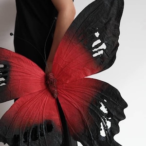 Puede incluir: Una gran mariposa decorativa con alas rojas y negras. Las alas son de un rojo vibrante que se desvanece en negro en las puntas, con una apariencia texturizada. La mariposa tiene un cuerpo y antenas detallados.