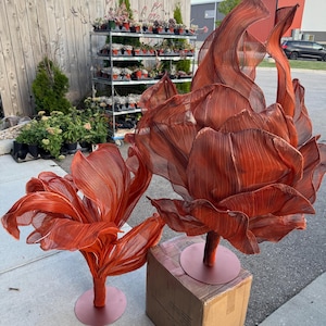 Può includere: Due grandi sculture floreali in tessuto arancione con un design strutturato e stratificato. I fiori sono montati su basi rotonde marrone-rossastre. Uno è su una scatola di cartone. Lo sfondo comprende una recinzione in legno e un portapiante in metallo.