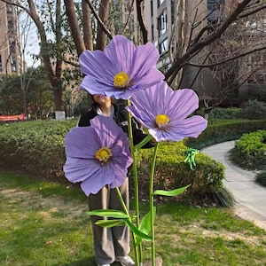Peut inclure: Trois grandes fleurs en papier violet avec des centres jaunes et des tiges vertes. Les fleurs sont expos&eacute;es &agrave; l'ext&eacute;rieur, debout dans une zone herbeuse. Les p&eacute;tales sont d'une teinte violette vibrante.