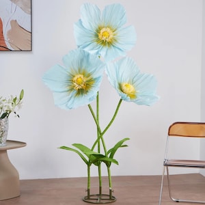 Peut inclure: Trois grandes fleurs de papier bleu clair avec des centres jaunes et des tiges vertes. Les fleurs artificielles ont des feuilles vertes &agrave; la base et sont dispos&eacute;es dans un pr&eacute;sentoir d&eacute;coratif. Les fleurs sont fix&eacute;es dans une base m&eacute;tallique.
