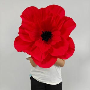 May include: A large, oversized decorative flower with vibrant red, ruffled petals and a textured, dark black center. The flower is constructed with multiple layers of petals that create a full, rounded shape, mimicking the appearance of a poppy.