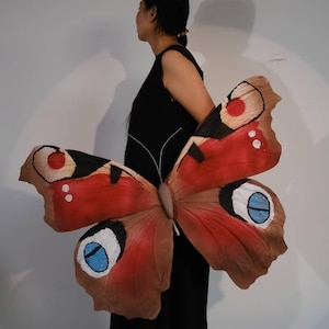 以下が含まれることがあります： 赤、茶色、黒、白のディテールが施された大きな蝶のコスチューム。 羽には、青と白のアクセントが付いた、特徴的な目の様な模様があります。 黒いドレスを着た人がコスチュームを着用しています。