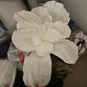 May include: A large white paper flower with multiple layers of petals. The flower is made of tissue paper and has a delicate, ruffled appearance.