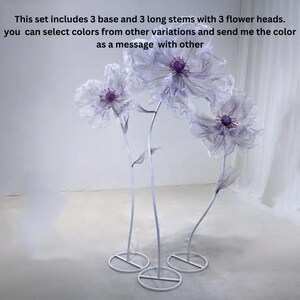 Puede incluir: Tres soportes de flores de metal blanco con tres grandes cabezas de flores blancas y transparentes con centros morados. Las cabezas de las flores est&aacute;n hechas de tela y est&aacute;n unidas a los soportes con tallos largos y blancos.