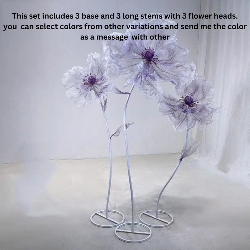 Puede incluir: Tres soportes de flores de metal blanco con tres grandes cabezas de flores blancas y transparentes con centros morados. Las cabezas de las flores est&aacute;n hechas de tela y est&aacute;n unidas a los soportes con tallos largos y blancos.