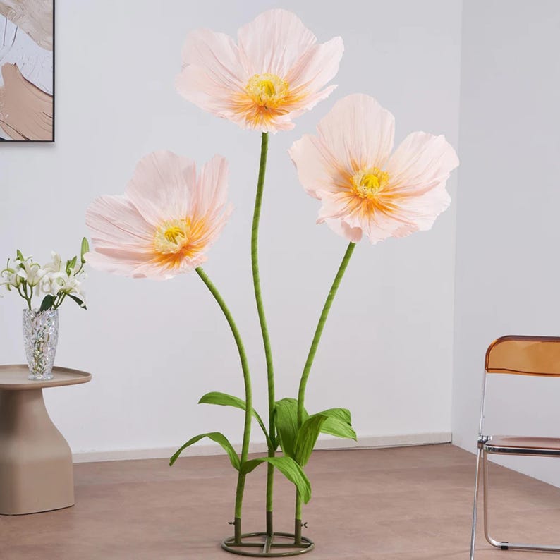 Peut inclure: Trois grandes fleurs en papier rose clair avec des centres jaunes et des tiges vertes. Les fleurs sont dispos&eacute;es dans une pr&eacute;sentation d&eacute;corative avec des feuilles vertes et une base m&eacute;tallique circulaire. Les tiges mesurent environ 152 cm de haut.