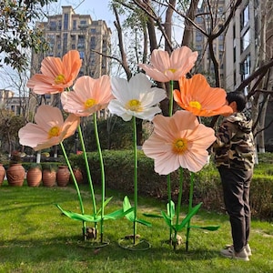Puede incluir: Grandes flores artificiales en tonos melocotón, blanco y naranja se alzan en una zona cubierta de hierba. Las flores tienen centros amarillos y tallos y hojas verdes. La escena es al aire libre, con edificios al fondo.