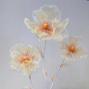 Puede incluir: Tres delicadas flores de papel blanco con centros naranjas. Las flores est&aacute;n dispuestas en tallos blancos delgados.