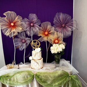 Puede incluir: Un pastel de boda de dos niveles decorado con detalles dorados y flores blancas, con un letrero de "Bride to Be" en la parte superior. El pastel est&aacute; sobre una mesa con un mantel blanco y tul verde. Hay cinco grandes flores de papel de seda de colores en el fondo.