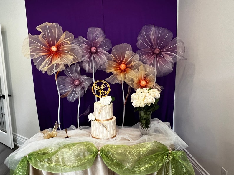 Puede incluir: Un pastel de boda de dos niveles decorado con detalles dorados y flores blancas, con un letrero de "Bride to Be" en la parte superior. El pastel est&aacute; sobre una mesa con un mantel blanco y tul verde. Hay cinco grandes flores de papel de seda de colores en el fondo.