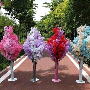 以下が含まれることがあります： ピンク、パープル、レッド、ブルーの花が咲いた人工の花の樹が4本。木は丸い土台が付いた金属製のスタンドの上に置かれています。