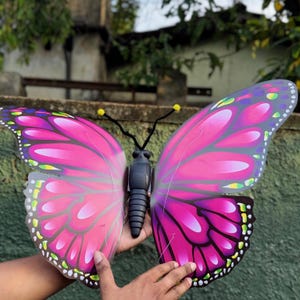 May include: A large, decorative butterfly with vibrant pink and purple wings. The butterfly has black antennae with yellow tips and a black body. The wings feature a detailed pattern with white, green, and black accents. The butterfly is held by hands.