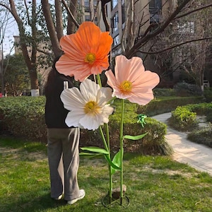Peut inclure: Trois grandes fleurs artificielles dans un jardin. Les fleurs sont orange, blanches et roses, avec des centres jaunes. Des tiges et des feuilles vertes soutiennent les fleurs. Les fleurs sont plac&eacute;es dans une base m&eacute;tallique circulaire.