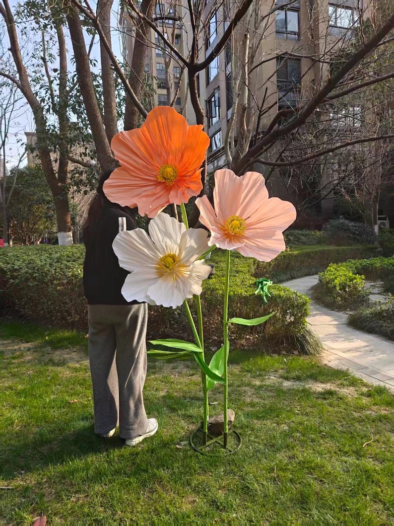 Peut inclure: Trois grandes fleurs artificielles dans un jardin. Les fleurs sont orange, blanches et roses, avec des centres jaunes. Des tiges et des feuilles vertes soutiennent les fleurs. Les fleurs sont plac&eacute;es dans une base m&eacute;tallique circulaire.