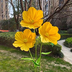 Peut inclure: Trois grandes fleurs artificielles jaune vif avec des tiges et des feuilles vertes. Les fleurs sont en pleine floraison, avec des p&eacute;tales d&eacute;taill&eacute;s et un aspect r&eacute;aliste. Les fleurs sont expos&eacute;es &agrave; l'ext&eacute;rieur dans une zone herbeuse.