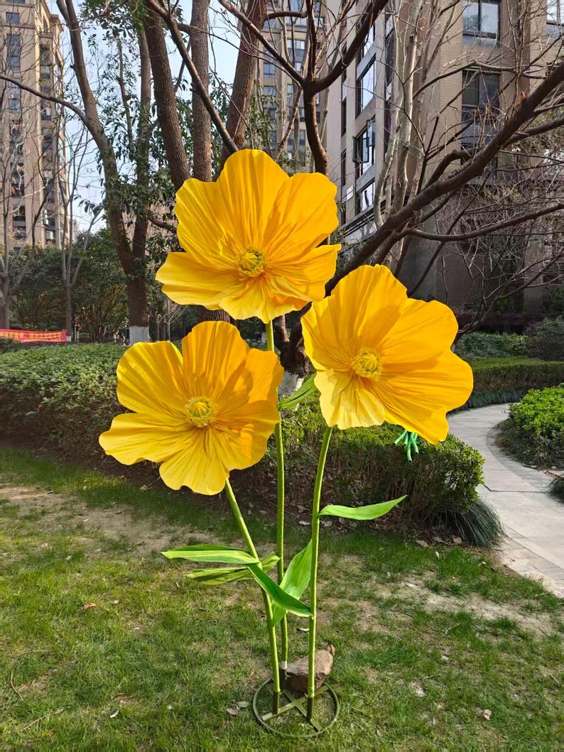 Peut inclure: Trois grandes fleurs artificielles jaune vif avec des tiges et des feuilles vertes. Les fleurs sont en pleine floraison, avec des p&eacute;tales d&eacute;taill&eacute;s et un aspect r&eacute;aliste. Les fleurs sont expos&eacute;es &agrave; l'ext&eacute;rieur dans une zone herbeuse.