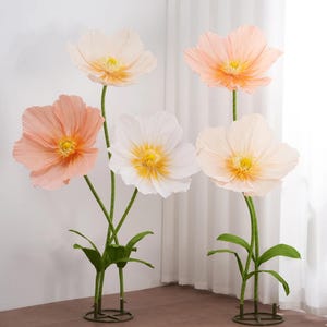 Peut inclure: Cinq grandes fleurs en papier aux tons p&ecirc;che, blanc et cr&egrave;me avec des centres jaunes. Les fleurs ont des tiges et des feuilles vertes et sont mont&eacute;es sur des supports m&eacute;talliques. Les fleurs sont dispos&eacute;es devant un rideau blanc.