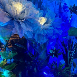 May include: A display of large, illuminated, blue and white fabric flowers. Lush green foliage and other plants are arranged around the flowers, creating a whimsical, dreamlike environment. The scene is bathed in blue light.