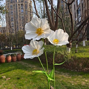Peut inclure: Trois grandes sculptures de fleurs blanches en m&eacute;tal avec des centres jaunes se dressent dans une zone herbeuse. Les fleurs ont des p&eacute;tales d&eacute;licats et froiss&eacute;s et sont soutenues par des tiges et des feuilles vertes. L'arri&egrave;re-plan comprend des arbres et un grand b&acirc;timent.